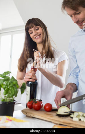 Mitte Erwachsene Frau Eröffnung Wein Flasche während reifer Mann schneiden Gemüse Lächeln Stockfoto