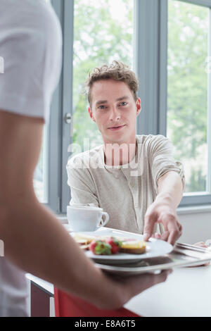 Junger Mann bietet Platte mit Frühstück mit einem anderen Mann, Lächeln Stockfoto