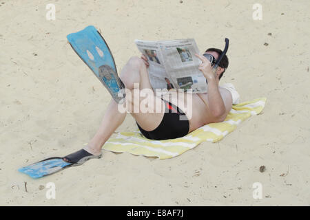 Mann in Flossen und Taucherbrille mit Schnorchel legt auf Sand und liest Zeitung Stockfoto