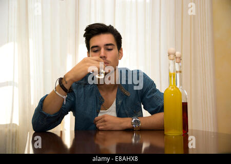 Junger Mann sitzen trinken allein an einem Tisch mit zwei Flaschen Schnaps neben ihm schlürfen von Shot-Glas, seinen Kummer zu ertränken Stockfoto