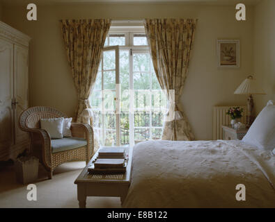 Zuckerrohr-Sessel neben französische Fenster mit floral gemusterte Vorhänge im Land Schlafzimmer mit Creme Bettwäsche Stockfoto