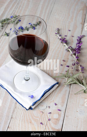 Ein Glas Rotwein auf einer Serviette und einem weißen rustikalen Holztisch. Blumen und Blütenblätter sind auf dem Tisch verstreut. Hochformat Stockfoto