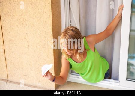 Kleine blonde kaukasische Mädchen mit Papierflieger im Fenster Stockfoto