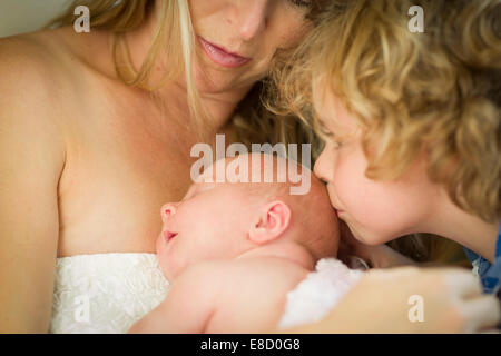 Schöne junge Mutter hält Neugeborene Baby Mädchen wie Bruder Küsse ihren Kopf. Stockfoto