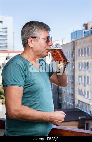 Ein älterer Mann trinkt Bier schlürfen es langsam mit Blick auf eine Industrielandschaft Stockfoto