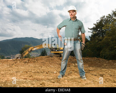 Kaukasische männlichen Ingenieur auf Baustelle mit Hammer, mit Bagger im Hintergrund stehend Stockfoto