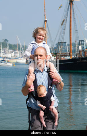 Reife Dad / Vater mit zwei / 2 Kinder; ein Kind auf Schultern und Baby im Tragetuch. Cornwall UK. Stockfoto