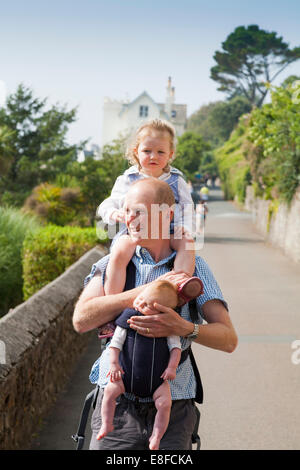 Reife Dad / Vater mit zwei / 2 Kinder; ein Kind auf Schultern und Baby im Tragetuch. Cornwall UK. Stockfoto