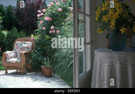 Gelbe Blumen in Vase auf den Tisch neben offenen Türen zur Terrasse mit einem geflochtenen Sessel neben rosa Kletterrosen Stockfoto