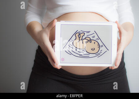 Schwangere Frau, die ein digitales Tablet mit einem Bild eines Babys vor ihrem Bauch hält Stockfoto