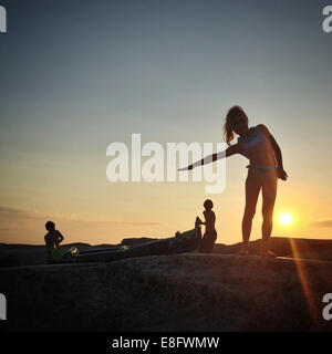 Silhouette von drei Kindern mit Kajak auf Felsen bei Sonnenuntergang, Norwegen Stockfoto