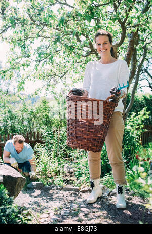 Porträt der glückliche Frau tragen Weidenkorb mit Mann Gartenarbeit im Hintergrund am Hof Stockfoto