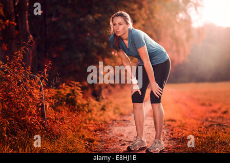 Junge sportliche Frau, die eine Pause vom Training stehen ruht ihr die Hände auf den Knien auf einem ländlichen Weg durch üppige Ackerland Stockfoto