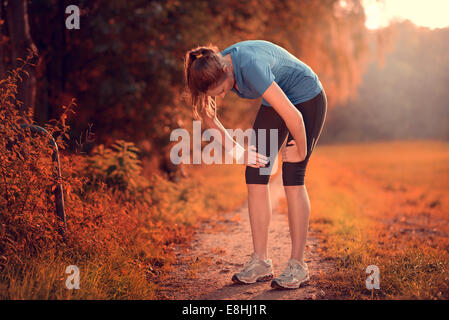 Junge sportliche Frau, die eine Pause vom Training stehen ruht ihr die Hände auf den Knien auf einem ländlichen Weg durch üppige Ackerland Stockfoto