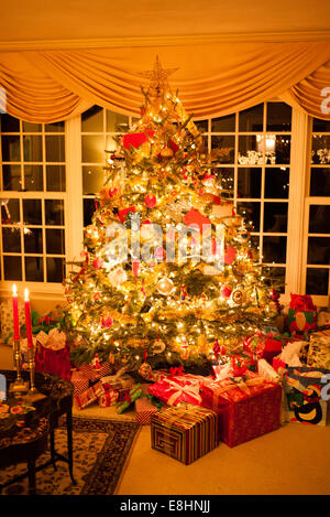 Weihnachtsbaum und Geschenke Vereinigte Staaten | VEREINIGTE STAATEN — Ein vollständig geschmückter Weihnachtsbaum mit einem Stern als Spitze steht vor drapierten Vorhängen, umgeben von verpackten Geschenken in einem Wohnzimmer. Die geschichteten goldenen Lametta, Glasornamente und gemischten Geschenkverpackungen spiegeln einen traditionellen amerikanischen Ansatz zur Feiertagsgeschenkgabe und zum Schmücken des Baumes wider. Stockfoto