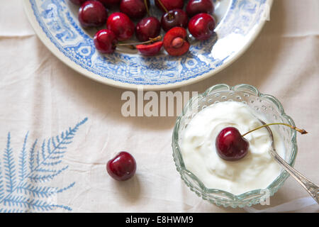 ganze Kirschen auf blaue Keramik-Platte, auf Blatt Leinen und Sahne oder Joghurt in Glasschale Stockfoto