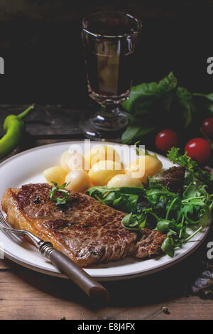 Gegrilltes Steak mit Kartoffeln und grünem Salat auf weißen Keramikplatte und Vintage Glas mit Rotwein über alten Holztisch. Stockfoto