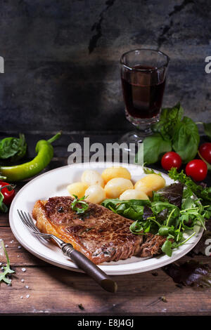 Gegrilltes Steak mit Kartoffeln und grünem Salat auf weißen Keramikplatte und Vintage Glas mit Rotwein über alten Holztisch. Stockfoto