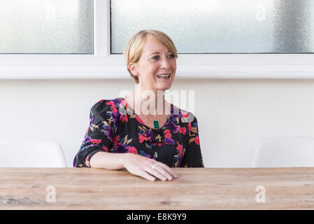 Eine Frau mit kurzen Haaren sitzt an einem Holztisch Lächeln auf den Lippen. Küche / Tagungsraum. Business-Frau / Mutter Stockfoto