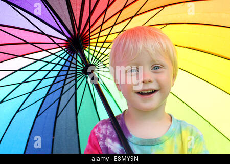 ein glückliches kleines zwei Jahre alten jungen Kind lächelt, als er draußen steht unter einem Regenbogen farbige Schirm vor dem Regen trocken zu halten Stockfoto