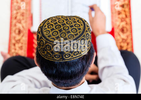 Asiatische muslimischen Mann liest Koran oder Koran auf beten Teppich tragen traditionelle Kleidung Stockfoto