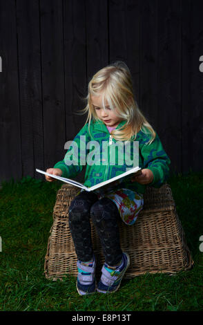 kleines Kind blondes Mädchen sitzen auf einem Weidenkorb ist das Lesen ein Buch im Freien, dunklen Holzwänden Hintergrund Stockfoto