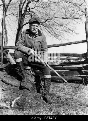 1940ER JAHRE ÄLTERER MANN SITZT AUF HOLZ SPLIT RAIL ZAUN HOLDING SCHUSS PISTOLE TRAGEN JAGDBEKLEIDUNG Stockfoto