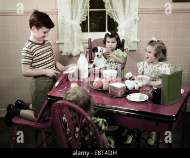 1940S 1950S BOY PEANUT BUTTER SANDWICH IN DER KÜCHE DREI MÄDCHEN UND HUND SITZEN HOCHSTUHL TISCH MIT ESSEN Stockfoto