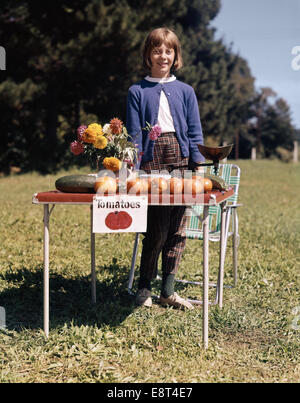 DER 1950ER JAHRE DER 1960ER JAHRE MÄDCHEN VERKAUFEN FRISCHE GARTEN TOMATEN MAIS ZUCCHINIBLÜTEN BLICK IN DIE KAMERA Stockfoto