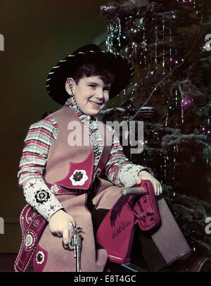 1940S 1950S IN COWBYO HUT KOSTÜM MIT SPIELZEUGPISTOLE HOLZPFERD WEIHNACHTSBAUM SITZEN LÄCHELNDE JUNGE GEKLEIDET Stockfoto