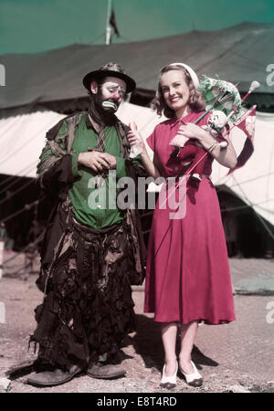 1940S 1950S LÄCHELNDEN BLONDEN FRAU IM CIRCUS HOLDING HOT DOG UND PREISE STEHEN NEBEN HUNGRIG TRAURIGER MANN HOBO CLOWN EMMETT KELLY Stockfoto