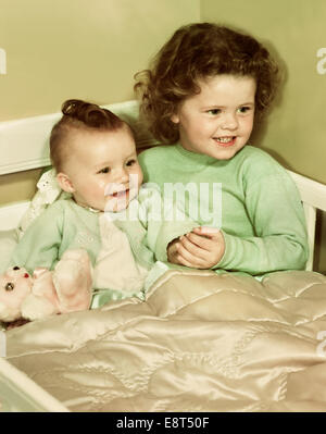 1940S 1950S LÄCHELNDE MÄDCHEN UND GLÜCKLICHES BABY SCHWESTER SITZEN ZUSAMMEN IN STÜCKCHEN KINDERBETT BETT Stockfoto