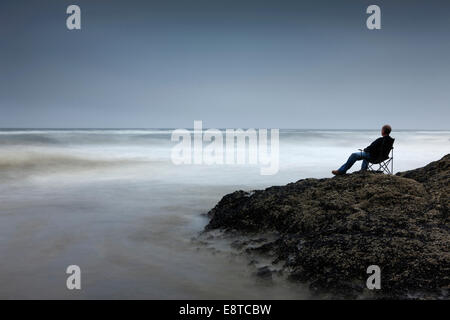 Kaukasischen Mann auf Stuhl mit Blick auf die Wellen am felsigen Strand Stockfoto