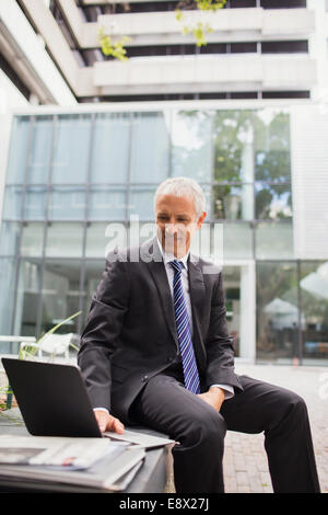 Geschäftsmann mit Laptop auf Bank außerhalb Bürogebäude Stockfoto