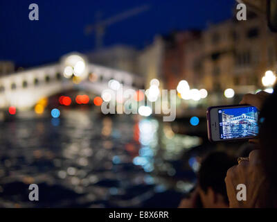 Frau auf einem Wasserbus nehmen ein Foto auf ihrem Smartphone der Rialto-Brücke in der Nacht in Venedig, Italien. Stockfoto