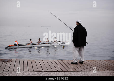 Thessaloniki, Griechenland. 17. Oktober 2014. Ein Mann Fische an der Küste von Thessaloniki als Ruderboot vor ihm, während des ersten Tages des Rudern Küsten-WM 2014 geht. Mehr als 350 Teilnehmer aus 23 Nationen nehmen Teil Credit: Giannis Papanikos/Alamy Live News Stockfoto