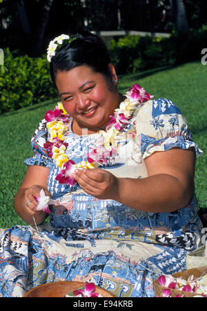 Eine lächelnde Frau Hawaii in Honolulu, Hawaii, USA, macht traditionellen Leis mit Plumeria (Frangipani) Blumen im Freien in der tropischen Sonne. Stockfoto