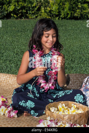 Eine 8 Jahre alte hawaiianische Mädchen nutzt eine lange Nadel Zeichenfolge Leis Orchidee und Plumeria Blumen im Freien in Honolulu, Hawaii, USA. Stockfoto