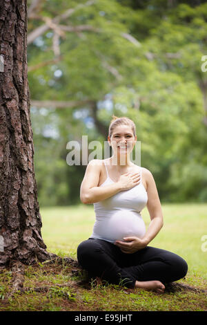 Porträt von weißen schöne schwangere Frau Yoga trainieren im Park in der Nähe von Baum, Herz und Bauch zu berühren Stockfoto