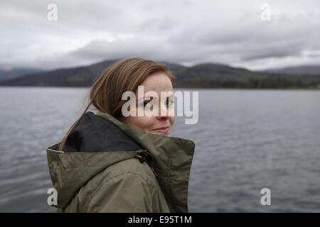 Hübsche junge Frau trägt einen Mantel steht lächelnd an den Ufern des Lake Windermere. Stockfoto