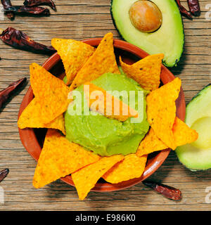 Nahaufnahme einer Schüssel Nachos mit Guacamole getaucht, auf einem Holztisch mit Avocado und Chili peppers Stockfoto