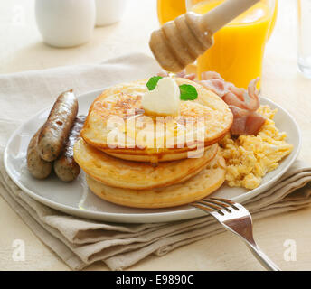 Großen herzhaftes Frühstück mit Würstchen, Eiern, Speck und einen Stapel von goldenen Pfannkuchen belegt mit einem nieselt Honig oder Sirup Stockfoto