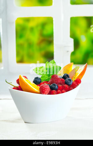 Schüssel mit frischen Beeren wie Himbeeren, Heidelbeeren und Johannisbeeren, garniert mit in Scheiben geschnittenen Orangen und Minze auf einem indoor Tisch mit Exemplar Stockfoto