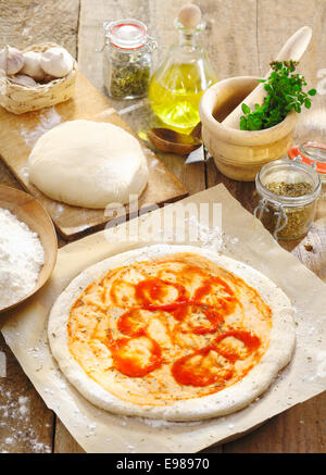 Vorbereitung eine italienische Pizza mit neu gerollt Tortekruste Basis mit Tomatenmark auf dem bemehlten Papier verteilt und umgeben von anderen Zutaten Stockfoto