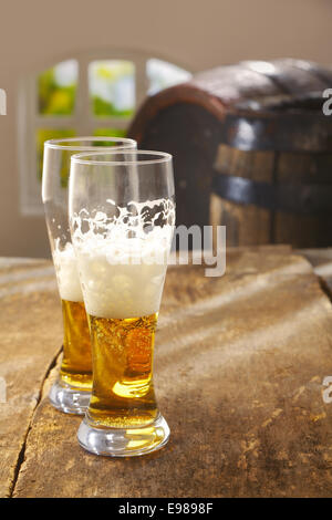 Zwei halb betrunken Gläser Bier mit einem guten schaumige Kopf stehen auf einem alten rustikalen Holztisch mit Vintage Holzfässern im Hintergrund Stockfoto