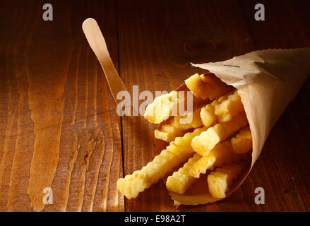 Crinkle cut goldene Pommes frites diente als ein Imbiss in einer Papierrolle braun liegend auf einem Holztisch mit Exemplar Stockfoto