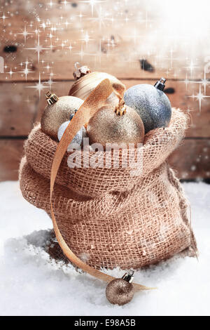 Dekorative Weihnachtskugeln in einer hessischen Tasche im Winterschnee hoch aufgetürmt und verziert mit einem goldenen Band inmitten tanzen funkelnden Sonnenlicht über eine Holzwand Stockfoto