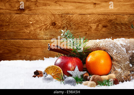 Künstlerische Weihnachten Stillleben mit buntem Obst und Gewürze aus der rustikale Jute-Tasche auf Schnee mit einem Holzbrett Hintergrund und Exemplar verschütten Stockfoto