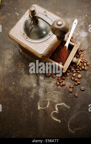 Draufsicht auf eine alte manuelle rustikale Holz Kaffeemühle mit einer Schublade voller frisch gemahlenen Kaffee mit verstreuten gerösteten Kaffeebohnen auf einem Grunge Schiefer mit Kritzeleien und Exemplar Stockfoto