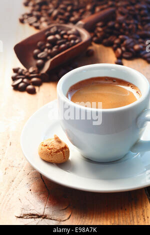 Morgendliche Tasse schaumig Espressokaffee auf einem Holztisch mit frisch gemahlenen gerösteten Kaffeebohnen gesehen im Hintergrund Verschütten von einer Kugel getroffen Stockfoto